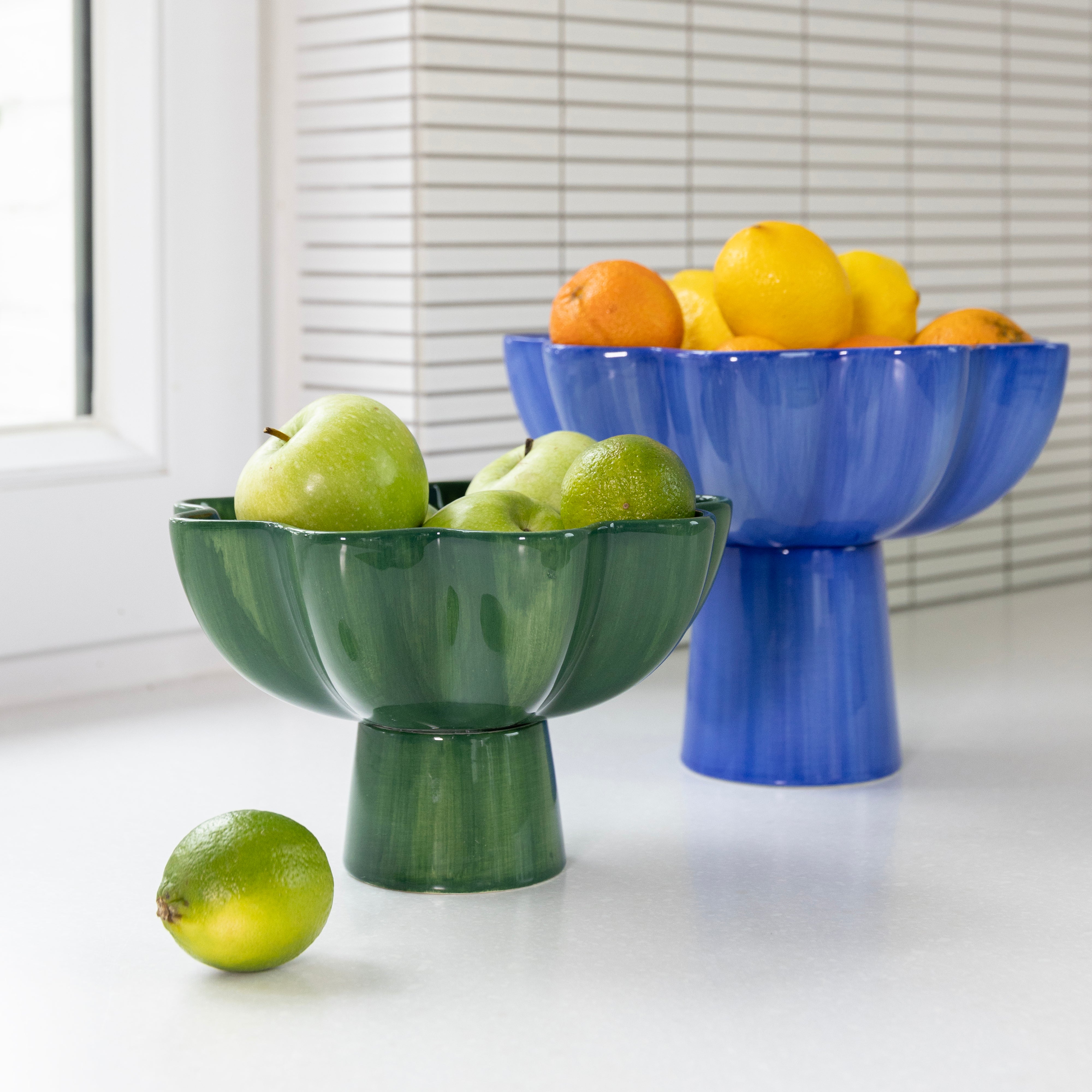Two decorative fruit bowls, one green and one blue, filled with fruits on a white surface. Also known as sun bowls